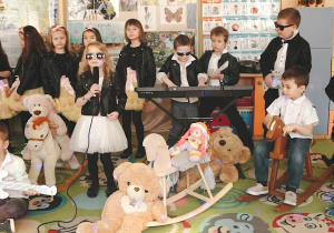 Przedszkolaki z grupy IV są przebrane w czerne ramoneski, trzech chłopców gra na gitarze, jeden na klawiszach, dwóch chłopców siedzi na koniakach na biegunach, dziewczynka z mikrofonem śpiewa, za nią stoją dziewczynki w chórku